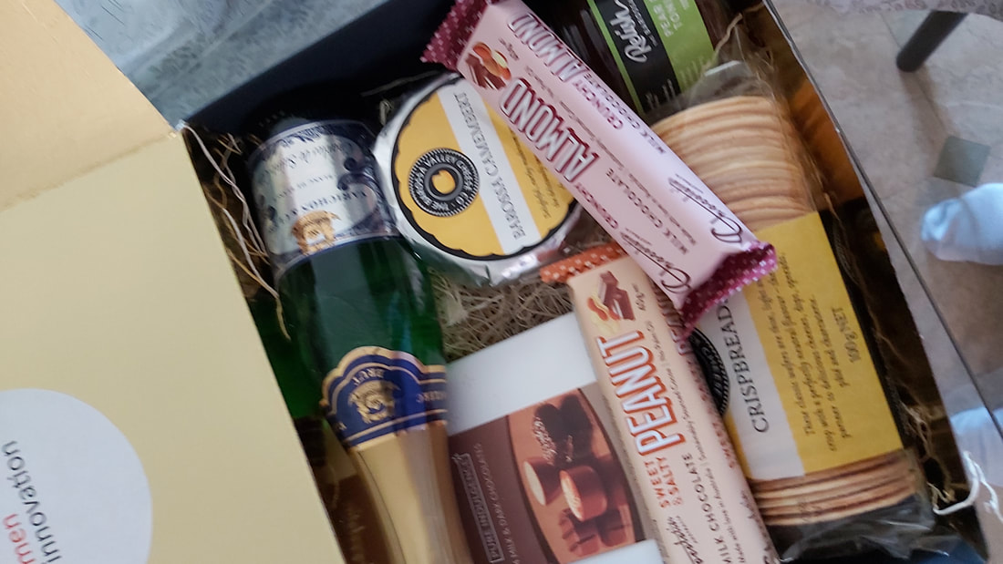 Opened gift box contains tiny champagne bottle, chocolates and chocolate bars, cheese, crackers and pear paste.