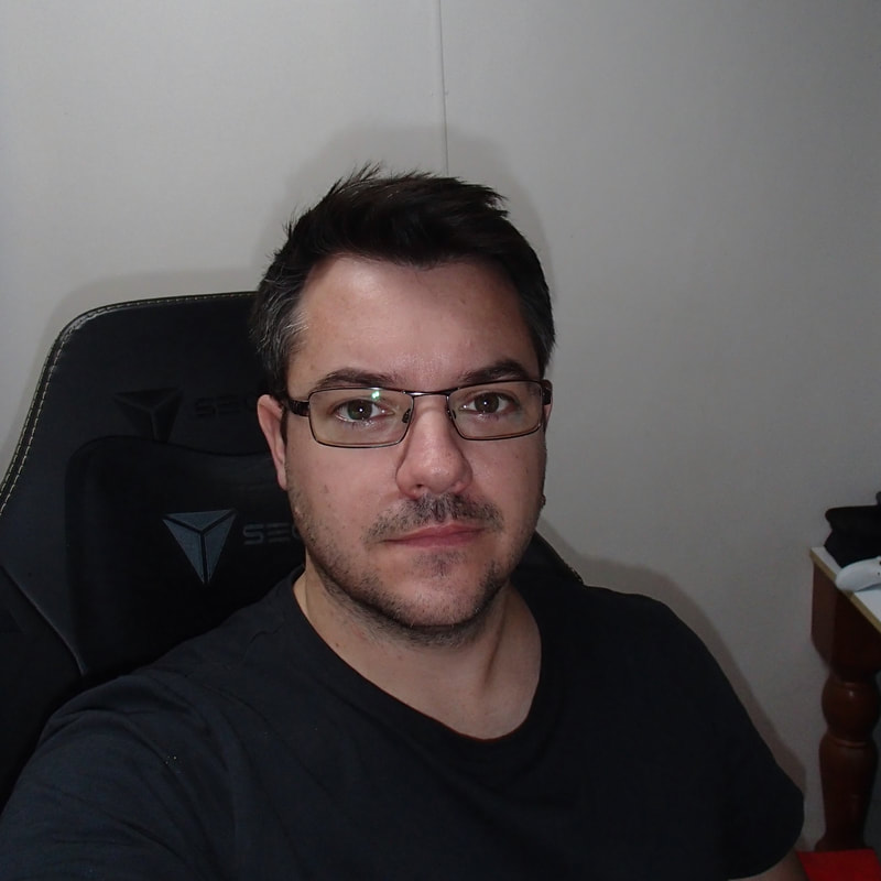 Head and shoulders of a handsome man in an office chair looking directly at the camera wearing glasses and sporting a moustache and the start of a beard.