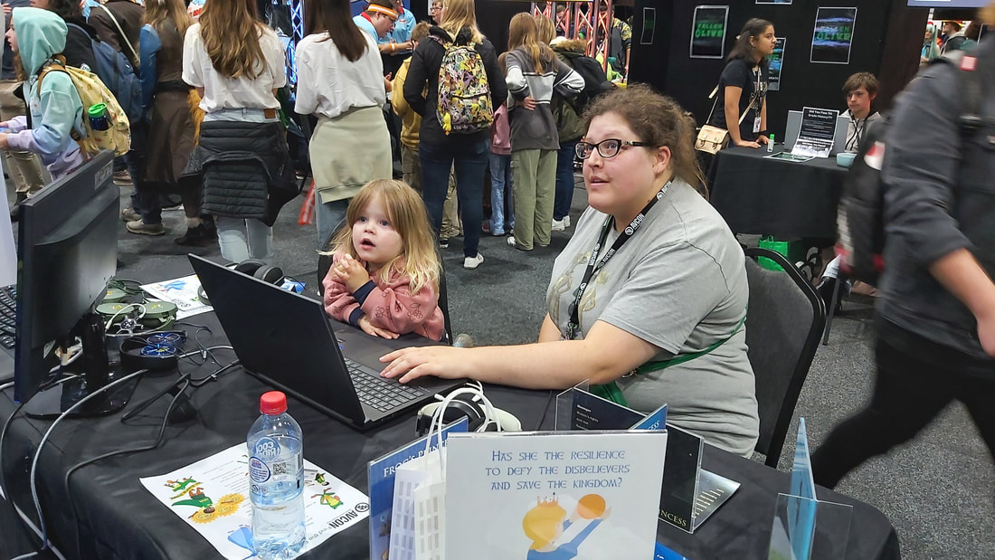 In a crowded convention centre, a mother and small girl sit at a booth.  The mother types on a keyboard and they both look up to watch the results off screen.  In the foreground is a table poster with an image of a princess saying Has she the resilience to defy the disbelievers and save the kingdom?