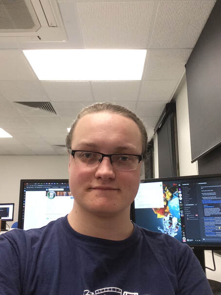 Picture of Max Winzar facing the camera with two monitors behind him.