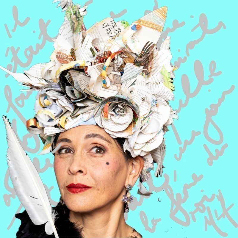 Headshot of Eliane Morel smiling wickedly wearing a tall headdress made of book pages curled into roses, feathers and sails, wearing a black feather boa and holding a white quill.