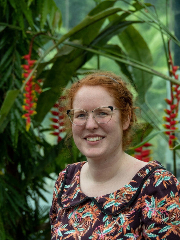 Head and shoulders shot of Louise Lawrence in front of palm fronds and red flowers which bring out the colour of her tied back hair.
