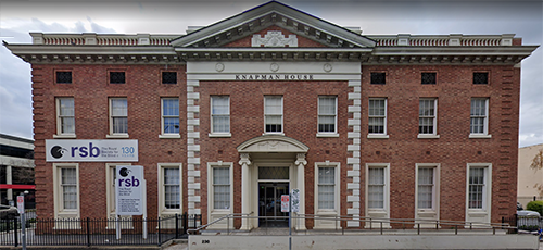 The imposing two-story brick building etched with a sign saying Knapman House, with a balcony on the roof and white painted window frames and portico, with RSB signs in front.
