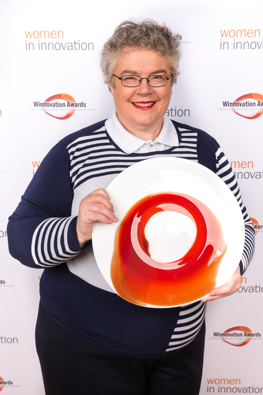 Kathy holds artistically painted plate in front of a display board decorated with Women In Innovation logos.